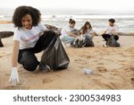 Positive young multiracial people volunteers in rubber gloves and african american lady with trash bag clean up garbage on sea beach, outdoor. Eco, environment conservation, protecting planet