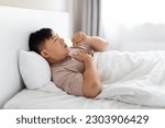 Small photo of Unhealthy sick middle aged asian man smoker wearing pajamas lying in bed and coughing, touching his chest, home interior, side view, copy space. Lung cancer, pneumonia, smokers morning cough