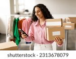 Small photo of Clothing Shop Entrepreneurship. Happy Lady Using Smartphone Holding Packed Cardboard Boxes With Clothes And Outfits, Sending Message To Buyer Standing In Store. Small Retail Business, Ecommerce