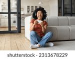 Excited Black Woman Playing Video Games On Smartphone At Home, Happy Cheerful African American Female Holding Mobile Phone And Celebrating Online Win While Sitting On Floor In Living Room, Copy Space
