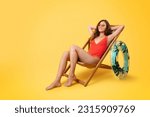 Cheerful millennial mixed race lady in swimsuit and sunglasses with inflatable ring sit in deck chair, enjoy vacation, isolated on yellow studio background. Holiday in summer, weekend and free time