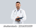 Small photo of Professional doctor man in medical coat with clipboard in hands posing over light grey wall background, middle aged physician with stethoscope looking at camera
