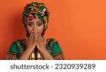 Small photo of Appalled emotional attractive young black woman wearing bright makeup and african costume covering her mouth, showing shock or astonishment, panorama, copy space, orange background