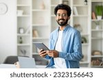 Small photo of Portrait Of Handsome Indian Male Freelancer With Digital Tablet In Hands Standing Near Desk At Home Office, Millennial Man Holding Tab Computer And Smiling At Camera, Enjoying Modern Technologies
