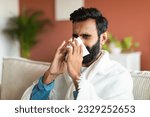 Small photo of Ill arabic guy sneezing and blowing runny nose in paper tissue sitting covered with blanket on sofa at home. Portrait of sick young man suffering from sinusitis illness, cold and influenza symptoms
