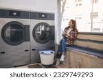 Young Woman In Casual Doing Laundry And Using Cell Phone, Sitting Waiting In Laundrette Room Near Washing Machines. Lady Booking Laundromat Via Mobile Application Posing Indoors