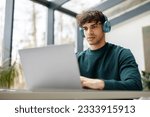 Small photo of E-learning concept. Focused european guy with wireless headphones looking at laptop, watching online tutorial, lecture or class sitting in university classrom or library