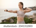 Small photo of Glad confident strong millennial caucasian woman athlete in sportswear doing arm exercises with dumbbells, enjoy workout, body care alone at sea beach. Sports, fitness outdoor