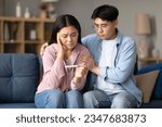 Small photo of Childbirth Problem. Depressed Korean Couple Holding Negative Pregnancy Test, Sitting Together On Couch At Home. Young Spouses Struggling With Infertility Issue And Dreaming About Baby
