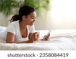 Nice App. Smiling Young Black Woman Using Smartphone And Drinking Coffee In Bed, Happy African American Female Relaxing With Mobile Phone In Bedroom, Browsing New Application, Copy Space