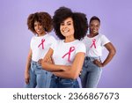Oncology Awareness Month. Group Of Three Black Ladies With Breast Cancer Ribbons On T Shirts Posing Together In Studio Over Purple Background, Looking At Camera. Selective Focus