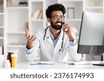 Small photo of Cheerful young Indian man physician, adeptly handling technology, converses on the phone while working on a computer at a clinic, symbolizing modern healthcare operations, copy space