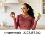 Small photo of Athletic woman in activewear choosing apple fruit or donut struggling from sugar cravings during diet, standing in kitchen setting. Unhealthy vs healthy food choice concept