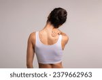 Small photo of Unrecognizable brunette woman with neck and back pain rubbing her painful body neck area, lady suffering from office syndrome, back view, grey studio background, copy space