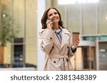 Happy attractive mature businesswoman in formal outfit beige trench coat have phone call, middle aged lady walking by street, talking on cell phone, drinking takeaway coffee, copy space