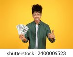 Small photo of Cashback, loan, giveaway. Rich wealthy excited handsome young black guy in casual outfit holding money cash dollar banknotes in his hand, showing thumb up over yellow studio background, copy space