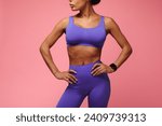 Cropped shot of fit black woman in purple sportswear standing with her hands on hips over pink studio background. Unrecognizable fitness model posing with tracker smartwatch