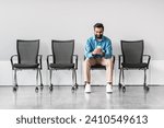 Casual indian businessman seated alone, absorbed in his smartphone, in stark waiting area with rows of empty chairs, suggesting moment of downtime