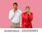 Astonished black couple covering mouths, man in white shirt and woman in red dress express surprise, set against pink background