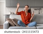 Small photo of Exuberant young woman in orange blouse enjoying music with headphones, singing joyfully while relaxing on a sofa in living room interior