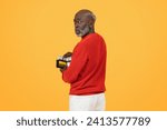 Small photo of Skeptical sad elderly African American man in a red sweater discreetly holding a wallet full of cash over his shoulder on a yellow background, looking wary or cautious