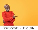 Small photo of An affable elderly glad gentleman in a bright red sweater with a beaming smile, playfully pointing to the side on a warm yellow background, exuding charm and friendliness