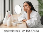 Small photo of Frustrated Indian Woman With Problem Skin Looking At Pimple On Cheek While Sitting In Front Of Mirror In Bathroom, Confused Eastern Lady In White Robe Suffering From Dull Skin, Closeup