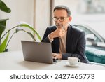 Small photo of Serious caucasian senior professional in a blazer intently analyzing information on a laptop screen with a hand on his chin, sitting at a white table with a coffee cup. Work, business