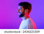 Profile portrait of young man with earbuds listening to online music, posing with eyes closed wearing wireless earphones on purple neon studio backdrop, enjoying his playlist