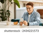 Small photo of Happy caucasian young man student with curly hair and glasses using his smartphone at a cafe table with a fresh croissant and coffee, enjoying a bright morning indoors. Devise for work