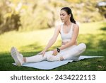 A caucasian millennial woman in white athletic wear gently stretches on a yoga mat in the park, her smartwatch visible as she focuses on her wellness routine, outside, full length