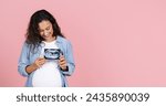 Small photo of Happy young brunette pregnant woman with big tummy showing ultrasound image of her baby, holding fetus photo over her chest, pink studio background, panorama with copy space