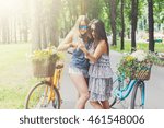 Happy boho chic stylish girls with mobile phone having fun. Beautiful women on bicycles with basket full of wild flower. Female friends, youth and happiness, active summer leisure in park concept.