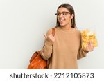 Small photo of Young student Indian woman holding crips isolated on white background points with thumb finger away, laughing and carefree.