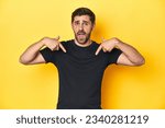 Small photo of Caucasian man in black t-shirt, yellow studio backdrop surprised pointing with finger, smiling broadly.