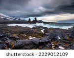 Waves crash onto the shoreline in front of Longdrangar, a pair of basalt rock outcrops on the Snaefellsnes Peninsula, Iceland. Volcanic rock and seaweed foreground with snow capped mountains beyond.