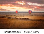 Sunrise over the Masai Mara, with a pair of low-flying hot air balloons and a herd of wildebeest below in the typical red oat grass of the region. In Kenya during the annual Great Migration.