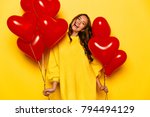 Young attractive girl with long curly hair, in yellow sweater holding red air balloons, posing at camera. St. Valentine's day. Isolated on yellow background.