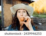 Small photo of Funny Woman Farmer in denim with a fresh egg, making a funny face. A woman in hat exhibits sheer happiness with a fresh egg in hand, standing on her farm Sustainable lifestyle, organic farmer