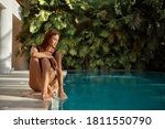 Attractive young woman kneeling by the edge of a swimming pool, touching the calm water with her hand