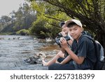 Asian boys wearing black t-shirt holding a binoculars sitting on stone by the river flowing down from the mountains in national park to observe fish in the river and birds on tree branches and on sky.