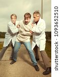 Small photo of Two older retro paramedic freak hunters in medical coats catching crazy man to straitjacket