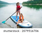 Children in swim life vest swimming on stand up paddle board together. Girls paddleboarding on SUP board on the lake. Active leisure with kids. Family local getaway concept