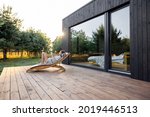 Young woman resting on sunbed and reading on a tablet on the wooden terrace near the modern house with panoramic windows near pine forest. Concept of solitude and recreation on nature