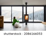 Small photo of Woman sitting with dog near fireplace and panoramic window at modern living room with stunning view on snowy mountains. Concept of rest in houses or cabins on nature. Idea of escape from everyday life