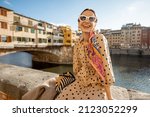 Young woman enjoys beautiful view on famous Old bridge in Florence, sitting on the riverside at sunset. Female traveler visiting italian landmarks. Stylish woman wearing dress and colorful shawl