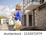 Small photo of Woman using phone while standing with mesh bag full of fresh vegetables and reusable coffee cup on porch of her house. Concept of sustainability, healthy food and modern eco-friendly lifestyle