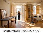 Small photo of Woman opens window blinds letting the sun inside the room, spending good morning in sunny and cozy apartment in beige tones. Interior view
