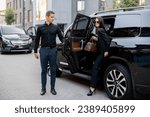 Small photo of Male driver helps a business lady to get out of a car, opening door of a luxury SUV taxi. Business lady with handbag wearing black formal wear. Concept of transportation service