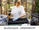 Small photo of Young woman filling seedling trays with a soil, sowing flower seeds at backyard. Tray with green sprouts in front. Concept of a hobby or small business of growing flowers.